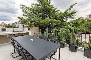 Terraza común ubicada en el quinto piso, cuenta con zona de asador de carbón y comedor exterior, así como sillas de estar. Disfruta la vista a la ciudad y sus hermosos atardeceres