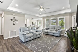 Bright living room with big screen TV, open kitchen & dining, flowing to a sunny backyard through large windows.