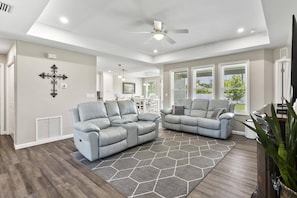 Bright living room with big screen TV, open kitchen & dining, flowing to a sunny backyard through large windows.