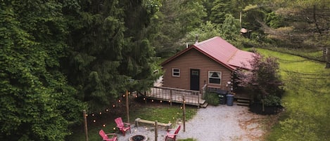 Charming forest cabin with a cozy fire pit surrounded by red chairs, perfect for evening gatherings. A serene natural retreat with lush greenery and warm outdoor lights to set the mood 🔥🌲✨