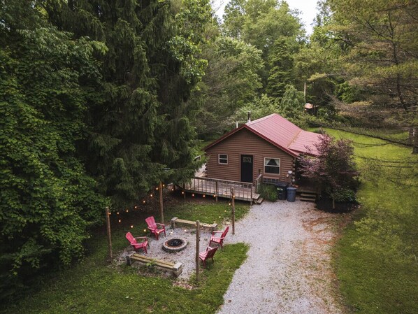 Charming forest cabin with a cozy fire pit surrounded by red chairs, perfect for evening gatherings. A serene natural retreat with lush greenery and warm outdoor lights to set the mood 🔥🌲✨