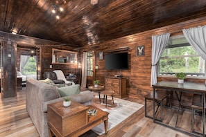 Relax in this rustic living room with rich wood tones, comfy seating, and a smart TV for cozy nights in 🪵📺. Large windows and warm lighting create a welcoming cabin vibe perfect for unwinding 🌲🛋️