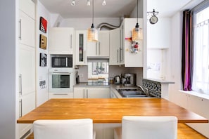 La pièce combine une cuisine ouverte et un espace repas chaleureux. Une table ronde en bois, des chaises modernes, un bar avec tabourets et des équipements complets. Une grande fenêtre créant une ambiance lumineuse et une pièce très conviviale.