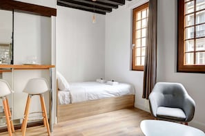 Un espace chaleureux et lumineux avec des touches de bois naturel et des fenêtres élégantes, offrant un coin nuit confortable, un coin repas moderne et un fauteuil accueillant pour se détendre.