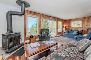 Cozy lakeside living room with wood-burning stove, rustic charm, and large windows offering stunning water views — the perfect spot to relax after a day of beach adventures!