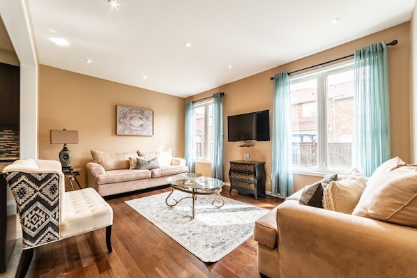Bright and cozy living room with elegant decor, large windows, and soft natural light. Features comfortable seating, a sleek wall-mounted TV, hardwood floors, and a stylish glass coffee table. Perfect for relaxing!