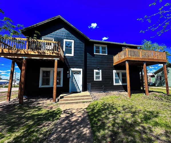 This striking house features bold black vinyl siding and elevated wooden decks, perfect for outdoor relaxation. Its unique design offers a modern aesthetic, complemented by a well-maintained lawn and inviting entryway.