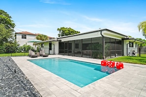 Beautiful backyard oasis features a sparkling swimming pool, comfortable lounge chairs, and lush tropical landscaping under a clear sky.