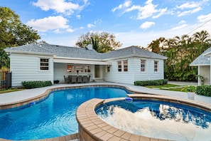 Beautiful backyard boasts a uniquely shaped swimming pool surrounded by lush greenery and a charming white home.