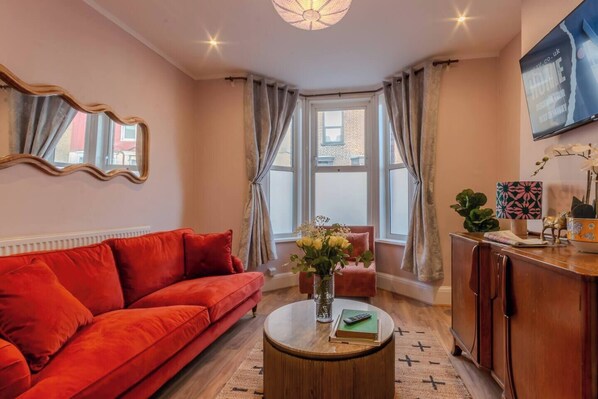 Stylish lounge with a bold orange sofa, smart TV, and Netflix — a cosy space to relax or work, filled with natural light from the bay window.