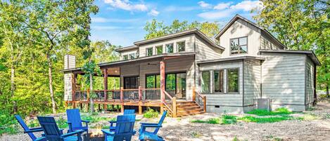 Contemporary forest home features spacious covered porch, blue Adirondack chairs around fire pit, and natural surroundings for ultimate outdoor living.