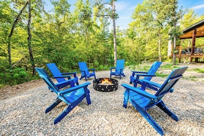 Sunlit forest clearing features vibrant blue Adirondack chairs encircling a star-studded fire pit for perfect evening gatherings.