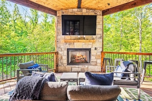 Cozy outdoor living room combines stone fireplace, mounted TV, and plush seating with breathtaking forest views for all-season enjoyment.