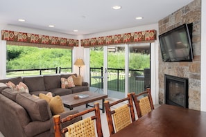 Cozy living area featuring a plush sectional sofa, stone fireplace with mounted TV, and expansive windows that open to a private patio.