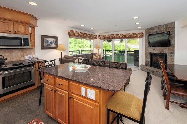 Inviting open-concept living space featuring a modern kitchen flowing seamlessly into a cozy living area with a sectional sofa, stone fireplace, and large windows that open to a wrap around patio.