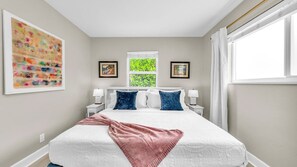 Unwind in a serene king bedroom featuring crisp white linens, plush velvet pillows, a smart TV, and cheerful coastal art. Your perfect El Cid retreat starts here.