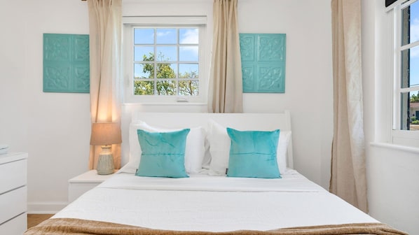 This breezy third bedroom features a queen bed with fresh white linens, coastal accent pillows, and a wall-mounted smart TV. Bright windows bring in natural light, creating a peaceful retreat perfect for relaxing after a day of exploring Palm Beach.