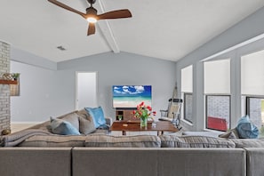 Spacious and sun-filled living room with vaulted ceilings, a large sectional sofa, and smart TV—perfect for movie nights or relaxing after a day of exploring. Enjoy natural light pouring in from the wall of windows and cozy up in a space designed for comfort and connection.