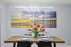 This modern dining area is both inviting and stylish, perfect for gathering with family or friends. A sleek wooden table is set for four, complete with contemporary black chairs and elegant tableware. A vibrant bouquet of red tulips adds a fresh and cheerful pop of color, tying in beautifully with the bold coastal artwork on the wall. The four-panel canvas captures a stunning s...