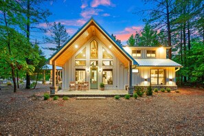 Welcome to our charming cottage - style retreat - glowing at twilight with a soft cream board and batten siding, a serene sage green door, cozy teak chairs for sipping morning coffee or tea.
