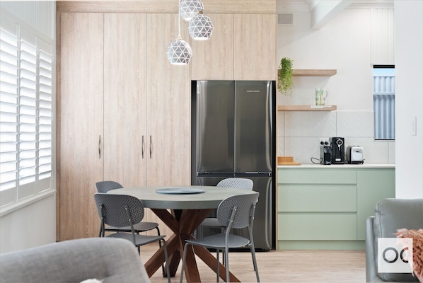 A modern kitchen and dining area with a sleek design. It features light wood cabinetry, a stainless steel refrigerator, a round dining table with perforated chairs, stylish geometric pendant lights, and light green lower cabinets. The space also includes 