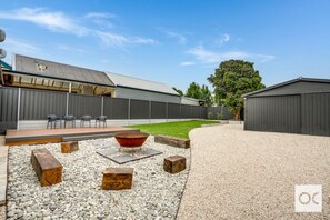 A well-organized backyard area with a gravel surface and a wooden deck. There are several wooden logs used as seating around a metal fire pit. In the background, there are metal sheds and a fenced area, with some greenery and trees adding to the serene at
