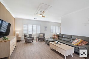 A modern and stylish living room with a light color scheme. It features a large leather sofa adorned with colorful cushions, two armchairs, a wooden TV stand, a wooden coffee table, and windows with shutters. The room has a wooden floor and a ceiling fan,