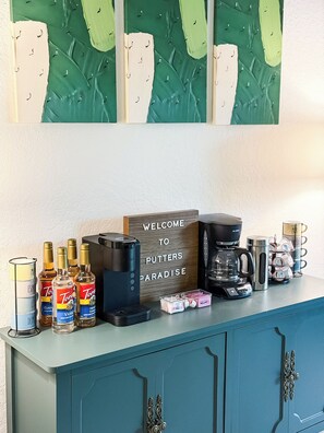 This dedicated coffee and beverage bar is a delightful amenity, offering a variety of options for guests. It features both a Keurig and a traditional drip coffee maker, an assortment of coffee syrups, and a welcoming "WELCOME TO PUTTERS PARADISE" sign, all set atop a stylish teal cabinet beneath abstract green and white artwork.