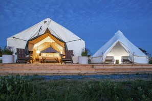 Two canvas tents lit at night with wooden deck and cozy chairs outside