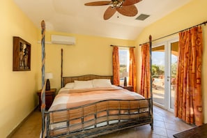 Cozy bedroom with a king-size bed, vibrant orange curtains, and natural light. Features unique artwork and a ceiling fan for added charm, with air conditioning for comfort. Ideal for a relaxing stay.