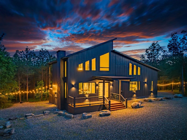 Modern cabin features dramatic architecture, warm lighting, and forest surroundings.