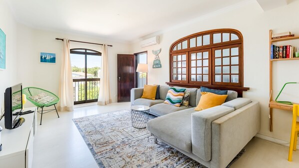 Bright, airy living room with cozy sofa, vibrant cushions, and large window letting in natural light. Modern decor with a relaxing vibe, perfect for unwinding after exploring. #LivingRoom #CozyStay #NaturalLight #ModernDecor #HolidayRental