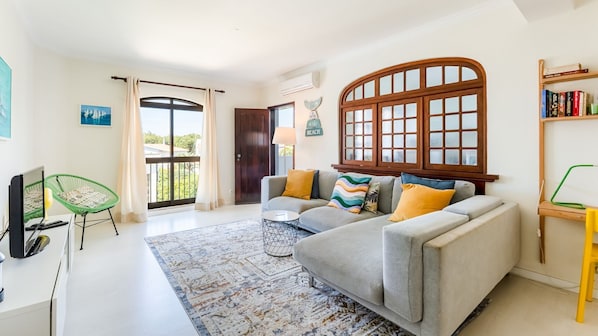 Bright, airy living room with cozy sofa, vibrant cushions, and large window letting in natural light. Modern decor with a relaxing vibe, perfect for unwinding after exploring. #LivingRoom #CozyStay #NaturalLight #ModernDecor #HolidayRental