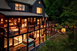 Experience luxury outdoor living on the spacious, illuminated decks of this stunning home, offering multiple areas for dining and lounging while surrounded by a serene wooded landscape. The warm glow of the exterior lights enhances the inviting ambiance fo