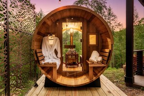 Indulge in a private wellness experience in this charming outdoor barrel sauna, featuring a warm wooden interior, a full glass door, and inviting bathrobes ready for relaxation. Nestled within a serene natural setting, it's the perfect escape for rejuvenat