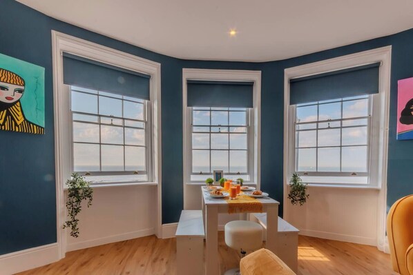 🍽 Dining with a View
Gather around the contemporary dining table beside stunning triple sash windows that frame the horizon—ideal for everything from relaxed breakfasts to sunset dinners.