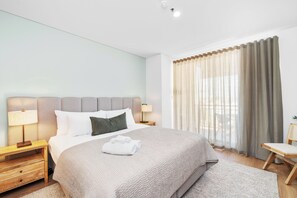 Unwind in this inviting bedroom, featuring a plush bed with a soft headboard, fresh white linens, and ample natural light filtered by elegant sheer and blackout curtains for a restful stay.