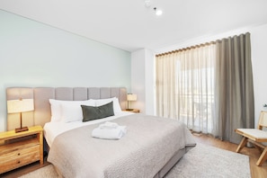 Unwind in this inviting bedroom, featuring a plush bed with a soft headboard, fresh white linens, and ample natural light filtered by elegant sheer and blackout curtains for a restful stay.
