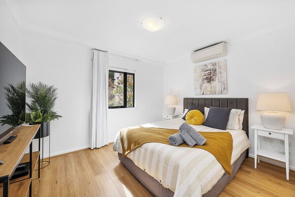 Bright, comfortable bedroom with a plush bed, air conditioning, and a TV. Features modern white and wood decor for a relaxing stay.