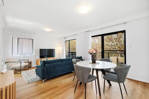 Bright open-plan living and dining area with a comfortable blue sofa, flat-screen TV, and a dining table for four. Features large windows for abundant natural light, stylish decor, and hardwood floors.