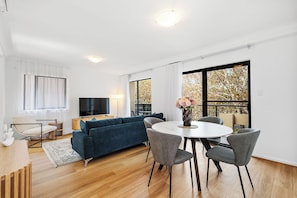 Bright open-plan living and dining area with a comfortable blue sofa, flat-screen TV, and a dining table for four. Features large windows for abundant natural light, stylish decor, and hardwood floors.