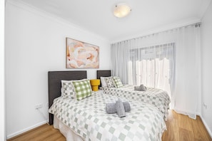 Bright bedroom with two cozy single beds, each featuring a green checkered duvet and accent pillows. The room is filled with natural light, thanks to the sheer curtains that allow it to stream in.