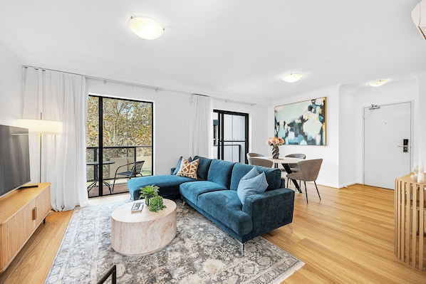 Spacious open-concept living and dining area featuring a comfortable blue sectional sofa, a flat-screen TV, and a dining table. Enjoy abundant natural light and balcony access in this stylish, modern space with hardwood floors.