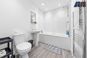 This bathroom is modern and fresh, with clean white finishes and stylish subway tiles around the bath. The wood-effect floor adds warmth, while the pedestal sink, mirror, and shelving cart keep it functional. The towel radiator is a nice finishing touch f