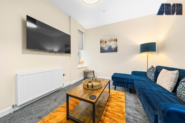 Vibrant and cozy lounge space featuring a plush navy sectional, warm lighting, and rustic wood accents — the perfect blend of comfort and style. With a large flat-screen TV, a bold orange rug, and thoughtful decorative touches, this room invites you to re