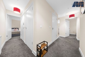Bright and spacious hallway designed for easy flow between rooms, featuring soft neutral walls, plush grey carpeting, and crisp white doors for a clean, modern feel. Pops of personality shine through with bold red ceiling light shades and a rustic wooden 