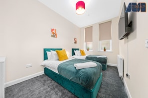 Inviting twin bedroom designed for comfort and style, featuring plush teal beds with crisp white linens and vibrant yellow accent pillows. Soft carpeting and natural light from dual windows create a cozy, restful atmosphere—complete with a wall-mounted TV