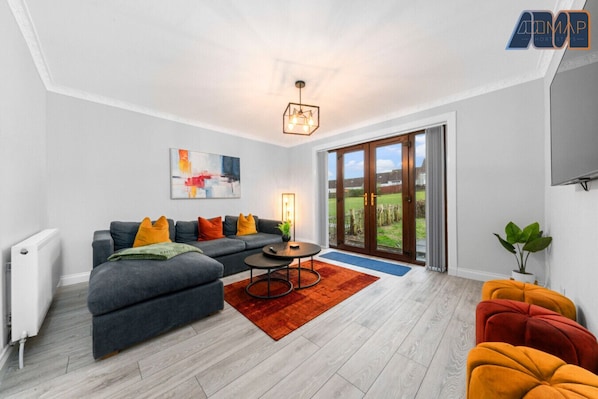 A stylish, cosy lounge with a spacious sofa, vibrant décor, and French doors leading to a serene outdoor view.