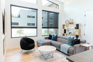 A cozy and bright living room with a comfy sofa, coffee table, and a TV. Light streams in through large windows, giving the space a warm, inviting feel.