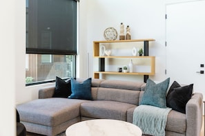 A cozy and bright living room with a comfy sofa, coffee table, and a TV. Light streams in through large windows, giving the space a warm, inviting feel.